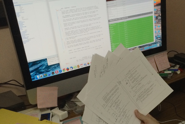 computer screen filled with scheduling charts and script breakdowns while a hand holds a heavily marked up script deep in pre-production.